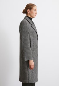Palton cu model houndstooth în alb și negru, cu guler îndoit, buzunare frontale și o siluetă lungă.