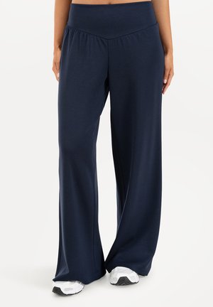 Persona que lleva pantalones anchos azul marino combinados con zapatillas blancas, de pie frente a un fondo liso y claro.
