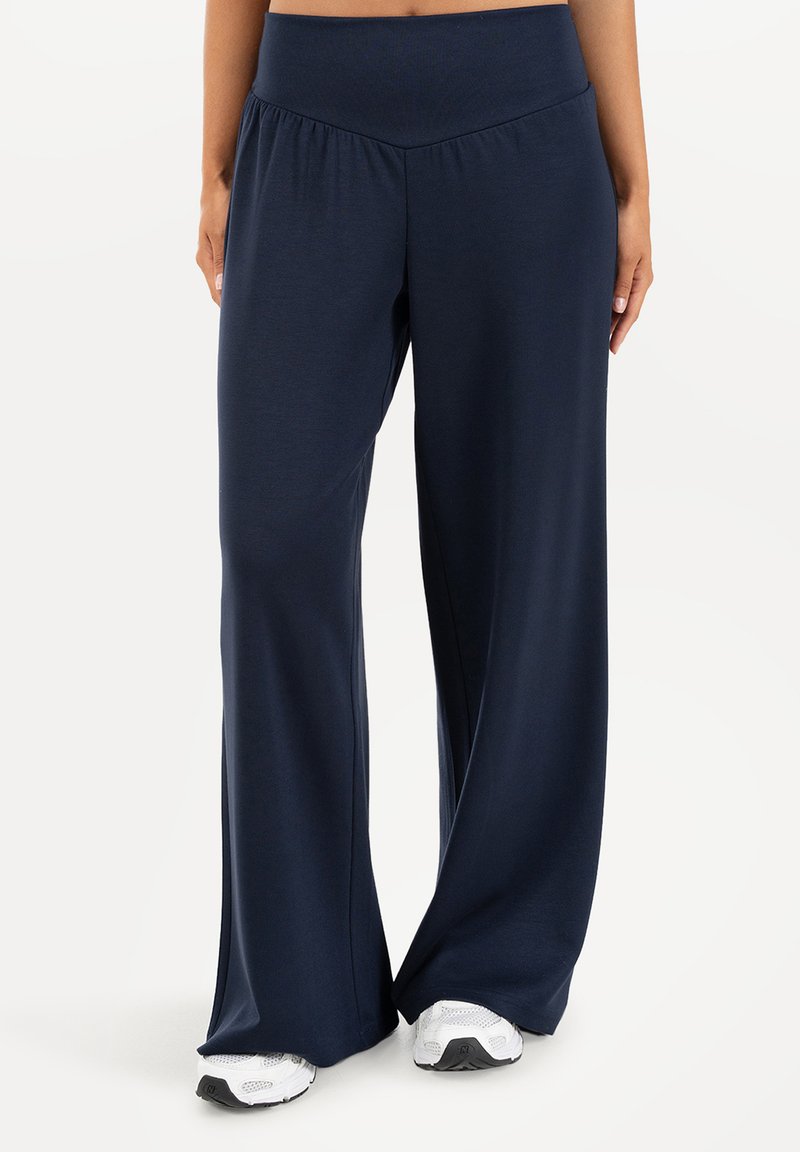 Personne portant un pantalon large bleu marine associé à des baskets blanches, debout devant un fond clair uni.