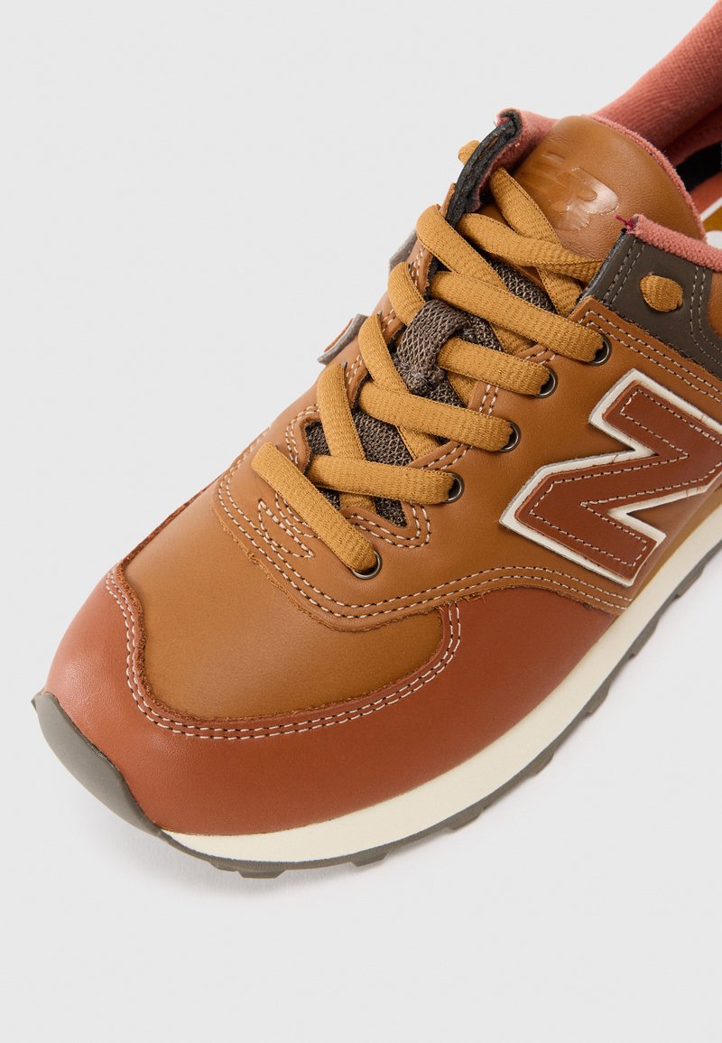 Brune lædersneakers med orange og grå snørebånd, hvid syning, gummisål og et fremtrædende "N" logo på siden.