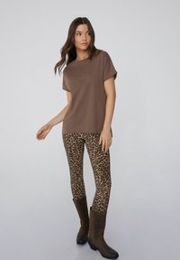 Brązowa koszulka z okrągłym dekoltem w połączeniu z legginsami w leopardzi wzór. Modelka nosi brązowe botki za kolano. Prosty, casualowy design z miękkimi teksturami.