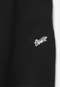 Tissu noir en gros plan avec logo texte brodé « SWITZ » en blanc près de la couture sur le côté droit.