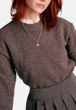 Brauner Strickpullover mit rundem Halsausschnitt und gerippten Bündchen. Kombiniert mit einem grauen, plissierten Rock und einer silbernen Kette mit Anhänger.