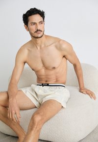 Beige shorts met een zwart-witte elastische tailleband, voorzien van een logo. Model zittend op een textuur-geïnspireerde, lichtgekleurde lounge stoel.