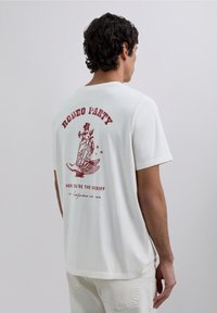 Camiseta de algodón blanca con un gráfico color burdeos que dice "RODEO PARTY" y un diseño alado en la parte posterior. Mangas cortas, ajuste relajado.
