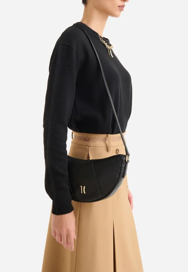 Donna che indossa un maglione nero e una gonna beige, con una piccola borsa a tracolla nera con logo dorato sul davanti.