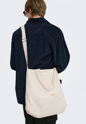Personne aux cheveux courts portant une chemise à carreaux foncée et des lunettes de soleil porte un grand sac fourre-tout en toile écrue avec bandoulière et poche avant.