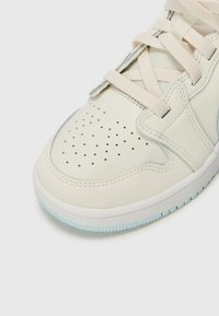 Zapatilla de color beige claro con caja de toe perforada, parte superior de cuero texturizado y sintético, cordones planos y una suela de goma azul translúcida.