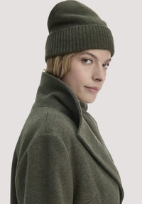 Olivgrüner Wollmantel mit hohem Kragen, kombiniert mit einer passenden gerippten Mütze. Weiche Textur, tailliertes Design, minimale Details.