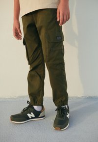 Olive green cargo pants with elastic cuffs, featuring multiple pockets. Paired with black sneakers with white and brown accents.