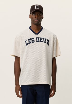 Cremefarbenes Netz-Jersey mit "LES DEUX" in dunkelblauer Schrift, V-Ausschnitt und kurzen Ärmeln, getragen zu dunkelblauen Hosen und einer braunen Mütze mit doppeltem "II".