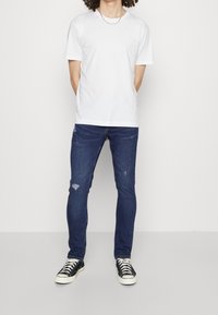 Camiseta de algodón blanca, vaqueros oscuros desgastados con ligeros desgarros y zapatillas negras con suelas blancas. Conjunto informal con un ajuste relajado.