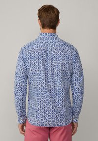Camisa de manga larga con una tela de patrón geométrico en azul y blanco, con cuello puntiagudo y puños abotonados. Se muestra la vista trasera.