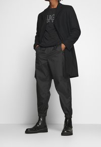 Musta pitkä takki mustan t-paidan päällä, jossa on valkoinen logo, yhdistettynä mustiin elastisiin housuihin ja mustiin nahkasaappaisiin.