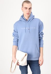 Lichtblauwe hoodie met een voorzak en een wit "TOMMY JEANS" logo. Gemaakt in combinatie met denim jeans en een lichtbeige tote bag.