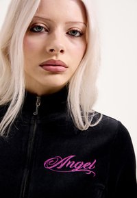 Puha anyagból készült fekete cipzáros dzseki, mellkasán rózsaszín "Angel" feliratú díszítéssel. Magas gallér és ezüst cipzár részlet.