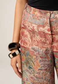 Pantaloni a gamba larga con motivo in morbido tessuto, caratterizzati da intricate fantasie floreali nei toni del rosso, blu e oro; accessoriati con bracciali chunky neri e crema.