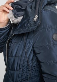 Detailní záběr osoby, která upravuje kapuci s kožešinovým lemem zamykací modré prošívané zimní bundy s viditelným stehem a nášivkou na rukávu.