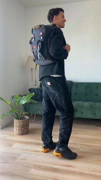 Black backpack with adjustable straps, featuring a front pocket and logo; worn over a black outfit, with orange accents on shoes.