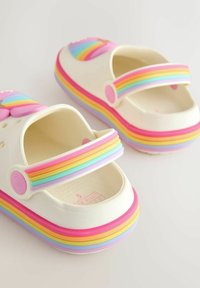 Valged kummist sussid mitmevärviliste vikerkaaretriipudega rihma ja tallal; neil on ventilatsiooniavad ja sile tekstuur.