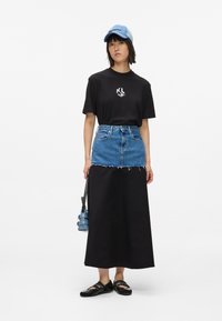 Fekete póló fehér logóval, ezüstkék farmer mini ruhával rétegezve, hosszú fekete szoknyával párosítva, hozzá egy kék sapka, fekete cipő és egy kis kék táska.