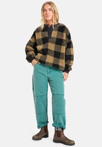 Vloze, zwart en tan geruite pullover met een zwarte kraag, gecombineerd met groene broek en bruine laarzen. Opvallende texturen en contrast in kleuren.