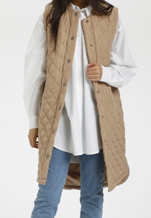 Beige gesteppte lange Weste mit Druckknöpfen. Kombiniert mit einem weißen Hemd, getragen über blauen Jeans. Hat ein Rautenmuster und eine glatte Textur.