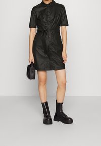 Czarna sukienka na krótki rękaw z kołnierzykiem, paskiem w talii i gładką teksturą. W połączeniu z masywnymi czarnymi botkami i małą czarną torebką.