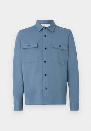 Mėlynas marškinėlis su sagomis, pagamintas iš lygaus audinio, turi apykaklę, dvi krūtinės kišenes su dangteliais ir juodas sagas priekyje.