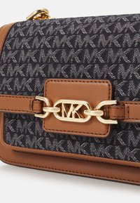 Brązowa skórzana torebka na klapę z niebieskim wzorzystym płótnem, złotymi akcesoriami oraz zapięciem w formie logo MK. Kompaktowy i strukturalny design.