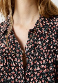 Blusa nera a fiori con piccoli fiori rosa e verdi, chiusura frontale con bottoni, e un tessuto morbido e leggero con una leggera lucentezza.