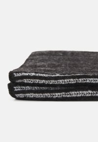 Coperta nera in lana piegata con una consistenza morbida, caratterizzata da un bordo cucito in bianco e grigio a contrasto. Forma rettangolare compatta.