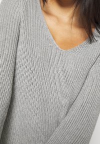 Pull gris en tricot avec un col en V et des manches longues. La texture est lâche et côtelée, mettant en avant un tissu doux et léger.