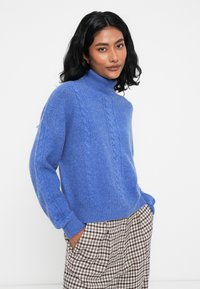 Modra pletena turtleneck pulover s teksturiranim vpletenim vzorcem. Ohlapen kroj, rahlo napihnjeni rokavi, v kombinaciji s karirastimi hlačami.