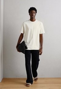 Tricou alb cu mânecă scurtă, pantaloni negri cu croială largă, ținând o geantă închisă. Încălțămintea are un design cu bretele. Siluetă simplă și curată.