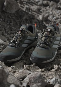 Gröna och svarta trailrunning-skor med snabblåsningssystem vilande på stenig terräng.