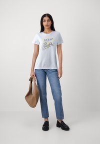 Camiseta de algodón blanca con el logo "GUESS" y un gráfico de triángulo amarillo, combinada con jeans azules y mocasines negros, sosteniendo una bolsa de mano marrón.