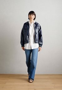 Jūras spēku faux ādas bumbvedēju jakas ar ribotiem piedurknēm un apakšmalu, valkāta virs baltas pogām aiztaisāmas krekla un gaiši ziliem platiem džinsiem. Gluda tekstūra.