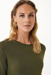 Zelený žebrovaný pletený top s vlnitým výstřihem. Modelka má vlnité blond vlasy a nosí malé zlaté kruhové náušnice. Detailní záběr.