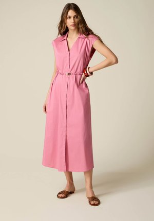 Vrouw draagt een mouwloze roze midi-jurk met ceintuur en kraag, gecombineerd met bruine sandalen en armbanden aan haar rechterpols.