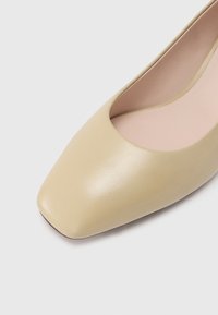 Beige leather pump with a square toe and a smooth texture. The interior is light pink, featuring subtle stitching along the edges.