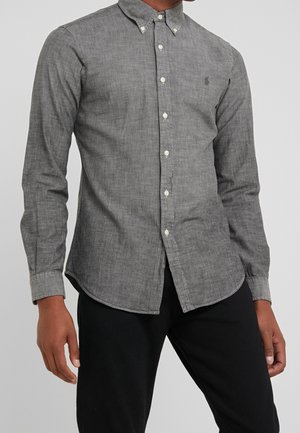 Homme portant une chemise grise à manches longues boutonnée avec un col boutonné et un pantalon noir, debout devant un fond uni.