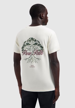 Mann trägt ein weißes T-Shirt mit einer Baumgrafik und den Texten "Pure Path" und "Rooted by friendship" auf dem Rücken vor einem einfarbigen Hintergrund.