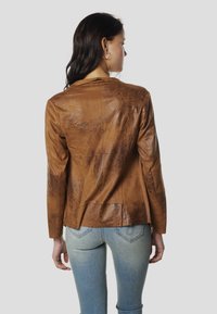 Jachetă din piele maro cu finisaj texturat, cu un design croită și mâneci lungi, purtată peste blugi albastru deschis.