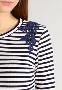 Haut à rayures à manches longues en tissu blanc et marine ; présente un détail de broderie florale marine sur l'épaule gauche. Encolure ronde.