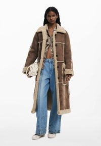 Cappotto in shearling marrone con una leggera fodera in pelliccia, vestibilità oversize, grande colletto e dettagli in bottone neri, indossato sopra un top a fantasia e jeans a gamba larga.