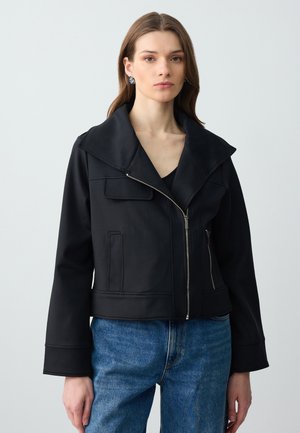 Giacca nera corta con ampio colletto, chiusura con zip, due tasche frontali e maniche strutturate, abbinata a jeans in denim blu.