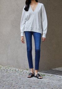 Sudadera gris de cuello en V con mangas largas, combinada con jeans ajustados de color azul oscuro y sandalias negras de tiras. Atuendo texturizado y casual.