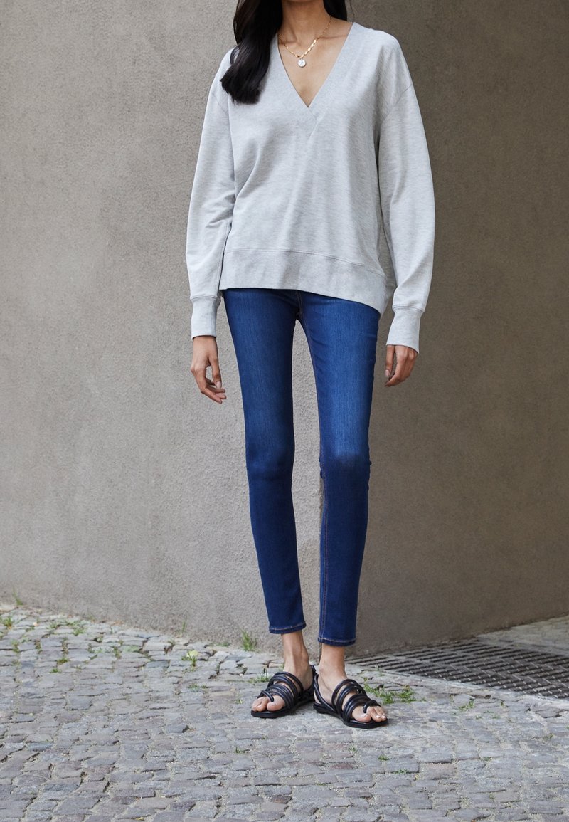 Sudadera gris de cuello en V con mangas largas, combinada con jeans ajustados de color azul oscuro y sandalias negras de tiras. Atuendo texturizado y casual.