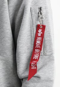 Felpa grigia con tasca con zip, caratterizzata da un'etichetta in tessuto rossa con la scritta "RIMUOVI PRIMA DEL VOLO" in bianco. Texture liscia.